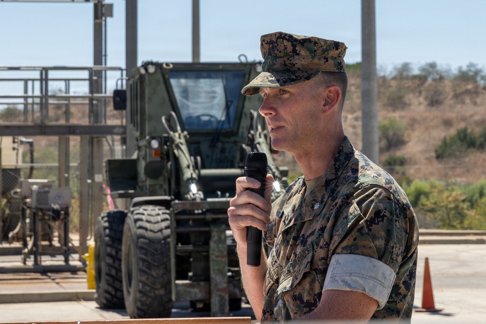 A U.S. Marine Corps officer in MARPAT digital camouflage uniform is speaking into a handheld microphone at an outdoor military facility. He appears to be addressing an audience at a formal event, possibly a ceremony or briefing. A military forklift or rough terrain vehicle is visible in the background.