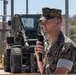 A U.S. Marine Corps officer in MARPAT digital camouflage uniform is speaking into a handheld microphone at an outdoor military facility. He appears to be addressing an audience at a formal event, possibly a ceremony or briefing. A military forklift or rough terrain vehicle is visible in the background.