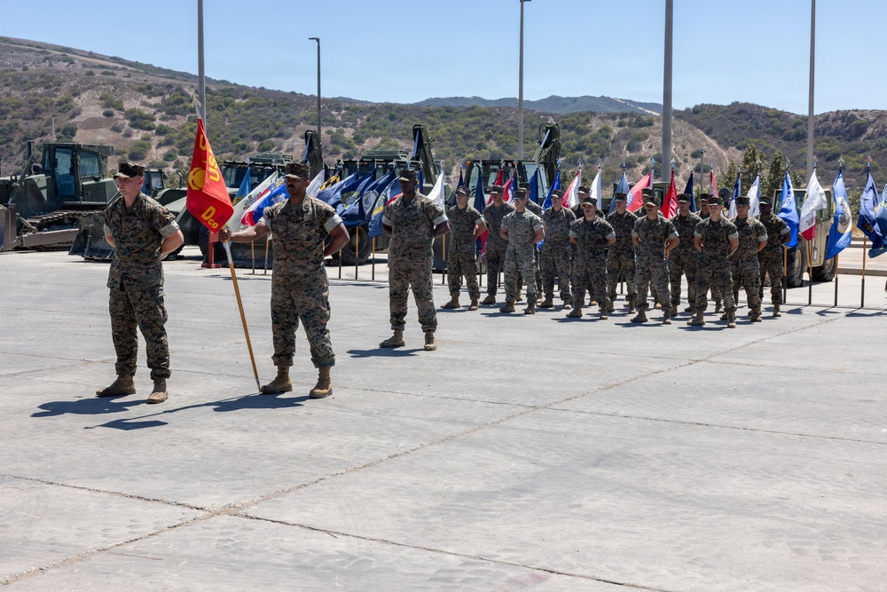 A military formation ceremony is taking place on a concrete parade ground, featuring personnel in MARPAT camouflage uniforms consistent with the United States Marine Corps. Multiple unit guidons and flags are visible being carried by flag bearers in a line formation. Heavy construction and engineering equipment is staged in the background, suggesting this may be a combat engineer or construction battalion unit.