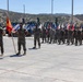 A military formation ceremony is taking place on a concrete parade ground, featuring personnel in MARPAT camouflage uniforms consistent with the United States Marine Corps. Multiple unit guidons and flags are visible being carried by flag bearers in a line formation. Heavy construction and engineering equipment is staged in the background, suggesting this may be a combat engineer or construction battalion unit.