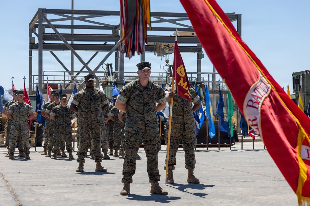 A formal U.S. Marine Corps military ceremony is taking place on a concrete parade ground or tarmac. Marines in woodland MARPAT camouflage uniforms are standing at parade rest in formation, with a color guard holding multiple unit guidons and flags. A steel construction framework and heavy equipment are visible in the background.