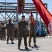A formal U.S. Marine Corps military ceremony is taking place on a concrete parade ground or tarmac. Marines in woodland MARPAT camouflage uniforms are standing at parade rest in formation, with a color guard holding multiple unit guidons and flags. A steel construction framework and heavy equipment are visible in the background.