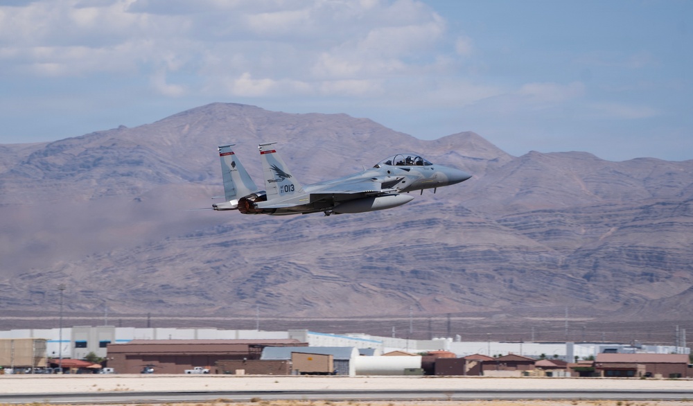 A gray F-15 Eagle fighter jet with tail number 013 is captured just after takeoff from a military airbase, with afterburners visibly engaged. The aircraft is climbing steeply against a backdrop of arid desert mountains and a partly cloudy sky. Military base infrastructure including hangars and buildings is visible in the background.