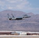 A gray F-15 Eagle fighter jet with tail number 013 is captured just after takeoff from a military airbase, with afterburners visibly engaged. The aircraft is climbing steeply against a backdrop of arid desert mountains and a partly cloudy sky. Military base infrastructure including hangars and buildings is visible in the background.