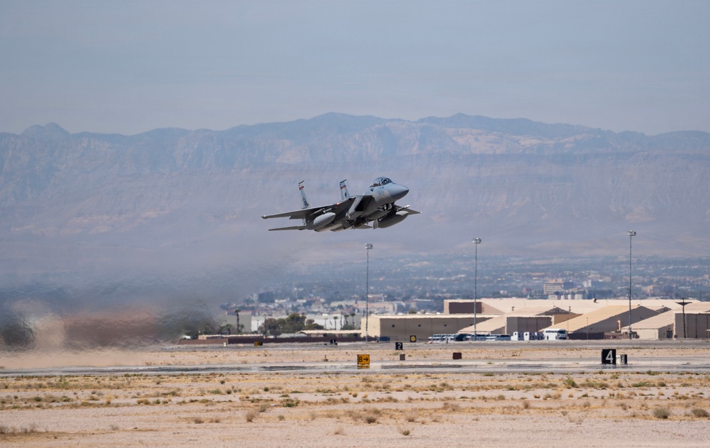 A military fighter jet, consistent with an F-15 Eagle design, is captured during takeoff or low-altitude flight over a desert airbase runway. The aircraft is in a steep climb attitude with landing gear still visible, suggesting a recent departure. The background reveals desert terrain, military hangars, and a mountain range under hazy atmospheric conditions.