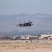A military fighter jet, consistent with an F-15 Eagle design, is captured during takeoff or low-altitude flight over a desert airbase runway. The aircraft is in a steep climb attitude with landing gear still visible, suggesting a recent departure. The background reveals desert terrain, military hangars, and a mountain range under hazy atmospheric conditions.