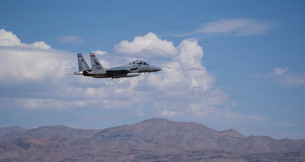 A military fighter jet, identifiable as an F-15 Eagle with tail number '017', is captured in flight against a partly cloudy blue sky. The aircraft is flying at low-to-medium altitude over an arid, mountainous desert landscape. The image appears to be taken during a training or operational flight in a desert region.