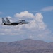 A military fighter jet, identifiable as an F-15 Eagle with tail number '017', is captured in flight against a partly cloudy blue sky. The aircraft is flying at low-to-medium altitude over an arid, mountainous desert landscape. The image appears to be taken during a training or operational flight in a desert region.