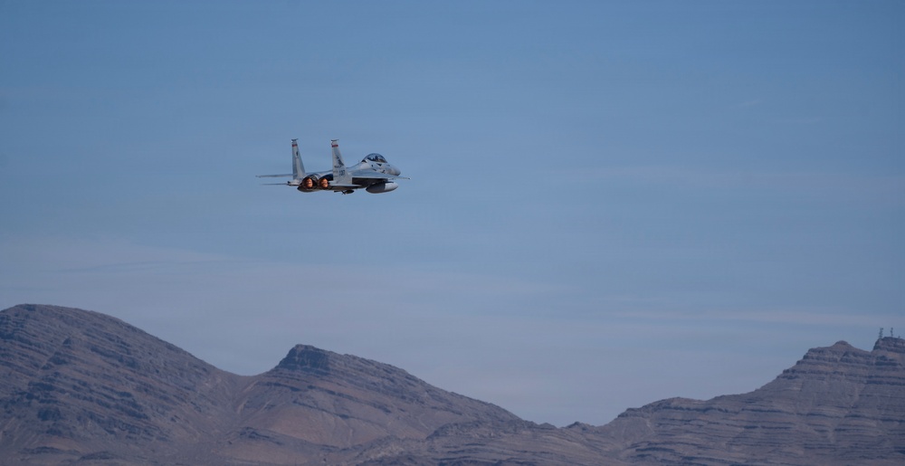 A military fighter jet, consistent with an F-15 Eagle, is captured in flight against a clear blue sky above a desert mountain range. The aircraft appears to be in level flight with afterburners visibly engaged, suggesting high-speed operation. The arid, rocky terrain below indicates a desert environment, possibly a military training range.