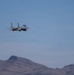 A military fighter jet, consistent with an F-15 Eagle, is captured in flight against a clear blue sky above a desert mountain range. The aircraft appears to be in level flight with afterburners visibly engaged, suggesting high-speed operation. The arid, rocky terrain below indicates a desert environment, possibly a military training range.