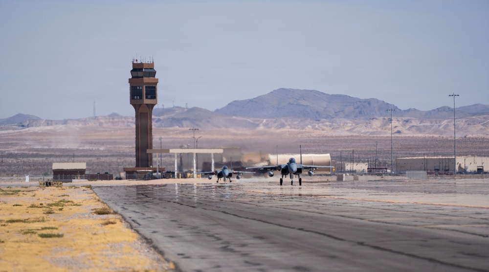 Two military jet aircraft are taxiing or taking off on a runway at a desert air base. A prominent air traffic control tower is visible in the background, along with various base infrastructure buildings and arid mountainous terrain. The setting appears to be a U.S. military installation in a desert environment, consistent with the American Southwest.