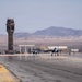 Two military jet aircraft are taxiing or taking off on a runway at a desert air base. A prominent air traffic control tower is visible in the background, along with various base infrastructure buildings and arid mountainous terrain. The setting appears to be a U.S. military installation in a desert environment, consistent with the American Southwest.