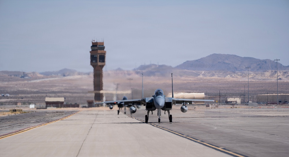 A military fighter jet, consistent with an F-15 Eagle, is taxiing on a runway at what appears to be a desert air base. A control tower is visible in the background along with arid terrain and mountains. The scene suggests routine flight operations at a military installation in an arid/desert environment.