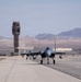 A military fighter jet, consistent with an F-15 Eagle, is taxiing on a runway at what appears to be a desert air base. A control tower is visible in the background along with arid terrain and mountains. The scene suggests routine flight operations at a military installation in an arid/desert environment.