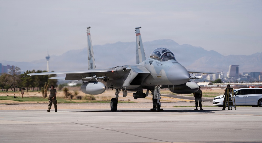 A U.S. Air Force F-15 Eagle fighter jet is taxiing or parked on a flight line at what appears to be Nellis Air Force Base near Las Vegas, Nevada. Ground crew personnel are positioned around the aircraft, likely performing post-flight or pre-flight checks. The Las Vegas Strip skyline and surrounding desert mountains are visible in the background.