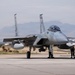 A U.S. Air Force F-15 Eagle fighter jet is taxiing or parked on a flight line at what appears to be Nellis Air Force Base near Las Vegas, Nevada. Ground crew personnel are positioned around the aircraft, likely performing post-flight or pre-flight checks. The Las Vegas Strip skyline and surrounding desert mountains are visible in the background.