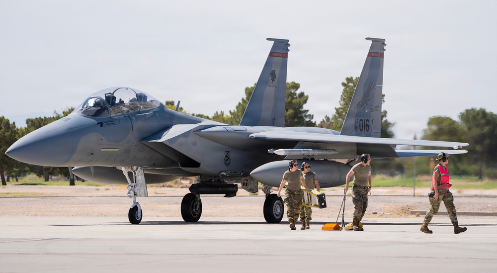 A U.S. Air Force F-15 Eagle fighter jet with tail number '016' is positioned on a flight line tarmac. Ground crew personnel in military uniforms and hearing protection are conducting pre- or post-flight maintenance and marshaling activities around the aircraft. The setting appears to be a desert air base, likely in the southwestern United States based on the vegetation and terrain.