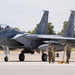 A U.S. Air Force F-15 Eagle fighter jet with tail number '016' is positioned on a flight line tarmac. Ground crew personnel in military uniforms and hearing protection are conducting pre- or post-flight maintenance and marshaling activities around the aircraft. The setting appears to be a desert air base, likely in the southwestern United States based on the vegetation and terrain.