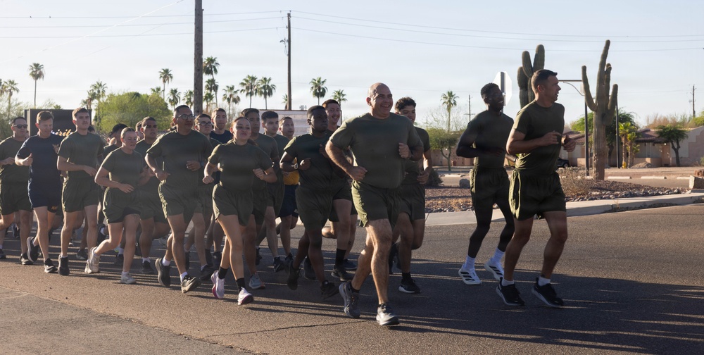A large group of military personnel in matching olive green t-shirts and shorts are conducting a formation run on a paved road. The setting appears to be a southwestern U.S. location, identifiable by saguaro cacti, palm trees, and desert landscaping in the background. The group is running in a tight formation, led by what appears to be a senior officer or NCO at the front.