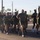 A large group of military personnel in matching olive green t-shirts and shorts are conducting a formation run on a paved road. The setting appears to be a southwestern U.S. location, identifiable by saguaro cacti, palm trees, and desert landscaping in the background. The group is running in a tight formation, led by what appears to be a senior officer or NCO at the front.