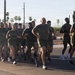 A large group of military personnel in matching olive green t-shirts and shorts are conducting a formation run on a paved road. The setting appears to be a southwestern U.S. location, identifiable by saguaro cacti, palm trees, and desert landscaping in the background. The group is running in a tight formation, led by what appears to be a senior officer or NCO at the front.