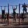 A group of military personnel in olive drab PT (physical training) uniforms are performing pull-ups on a red outdoor pull-up bar station. Three individuals are actively on the bars while others stand nearby observing or waiting their turn. The setting appears to be an outdoor fitness area on a military installation, likely during a physical fitness test or training session.