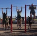 A group of military personnel in olive drab PT (physical training) uniforms are performing pull-ups on a red outdoor pull-up bar station. Three individuals are actively on the bars while others stand nearby observing or waiting their turn. The setting appears to be an outdoor fitness area on a military installation, likely during a physical fitness test or training session.