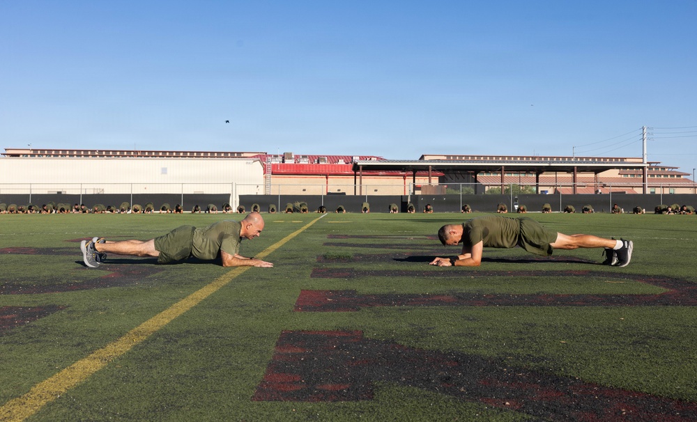 Two individuals in matching olive/military green t-shirts and shorts are performing push-ups or a low plank exercise on an artificial turf athletic field. In the background, a large group of similarly dressed individuals can be seen in a similar exercise position along the far end of the field. The setting appears to be a military or institutional athletic facility with stadium-style structures visible in the background.