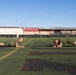 Two individuals in matching olive/military green t-shirts and shorts are performing push-ups or a low plank exercise on an artificial turf athletic field. In the background, a large group of similarly dressed individuals can be seen in a similar exercise position along the far end of the field. The setting appears to be a military or institutional athletic facility with stadium-style structures visible in the background.