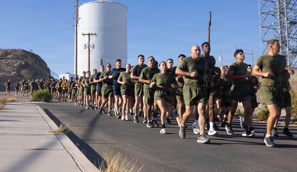 A large formation of military personnel, likely U.S. Marines based on their olive green PT (physical training) uniforms, are conducting a group formation run along a paved road. The run appears to take place at a military installation in an arid, desert environment. The group spans dozens of individuals running in a wide, organized formation.