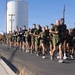 A large formation of military personnel, likely U.S. Marines based on their olive green PT (physical training) uniforms, are conducting a group formation run along a paved road. The run appears to take place at a military installation in an arid, desert environment. The group spans dozens of individuals running in a wide, organized formation.