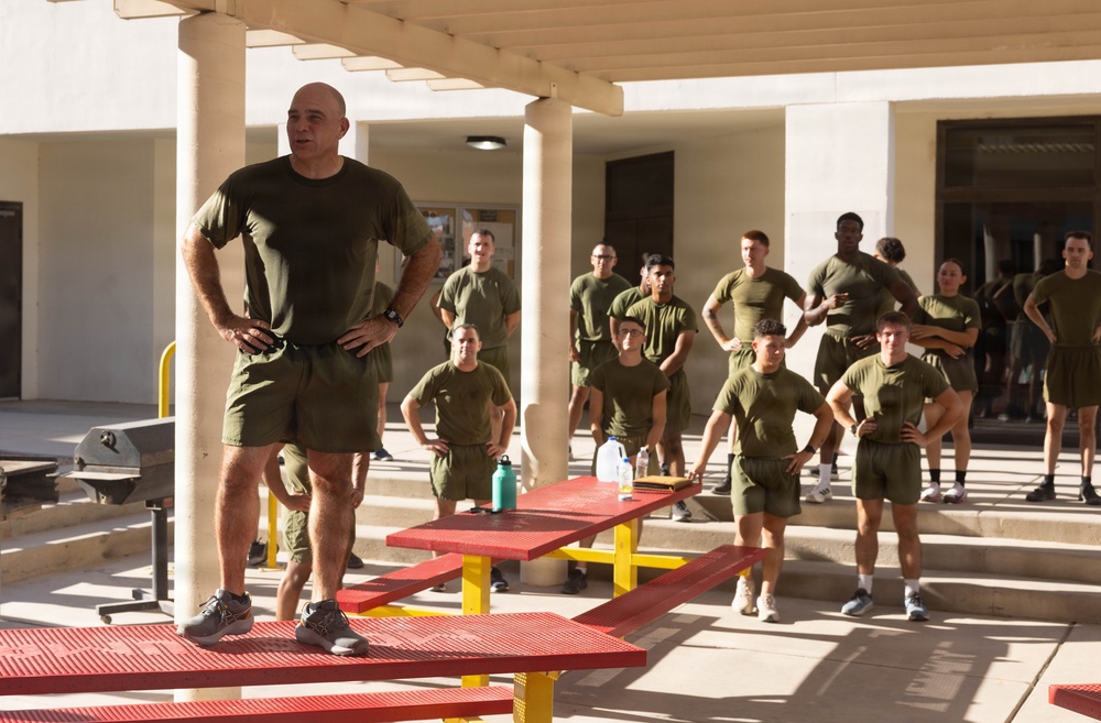 A senior military figure, dressed in olive green physical training (PT) uniform, stands elevated on a red picnic table addressing a group of uniformed personnel gathered in an outdoor courtyard. The group, also wearing matching olive green PT uniforms, appears to be listening attentively in a formation-like arrangement. The setting appears to be a military installation or base facility.