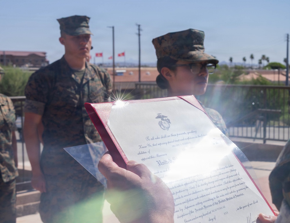 Two U.S. Marines in woodland MARPAT camouflage uniforms are participating in what appears to be a military promotion or appointment ceremony outdoors. A document bearing the United States Marine Corps Eagle, Globe, and Anchor emblem is being held up, consistent with a military appointment or promotion certificate. The setting appears to be a military installation, likely in a desert region based on the arid landscape and palm trees visible in the background.