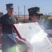 Two U.S. Marines in woodland MARPAT camouflage uniforms are participating in what appears to be a military promotion or appointment ceremony outdoors. A document bearing the United States Marine Corps Eagle, Globe, and Anchor emblem is being held up, consistent with a military appointment or promotion certificate. The setting appears to be a military installation, likely in a desert region based on the arid landscape and palm trees visible in the background.