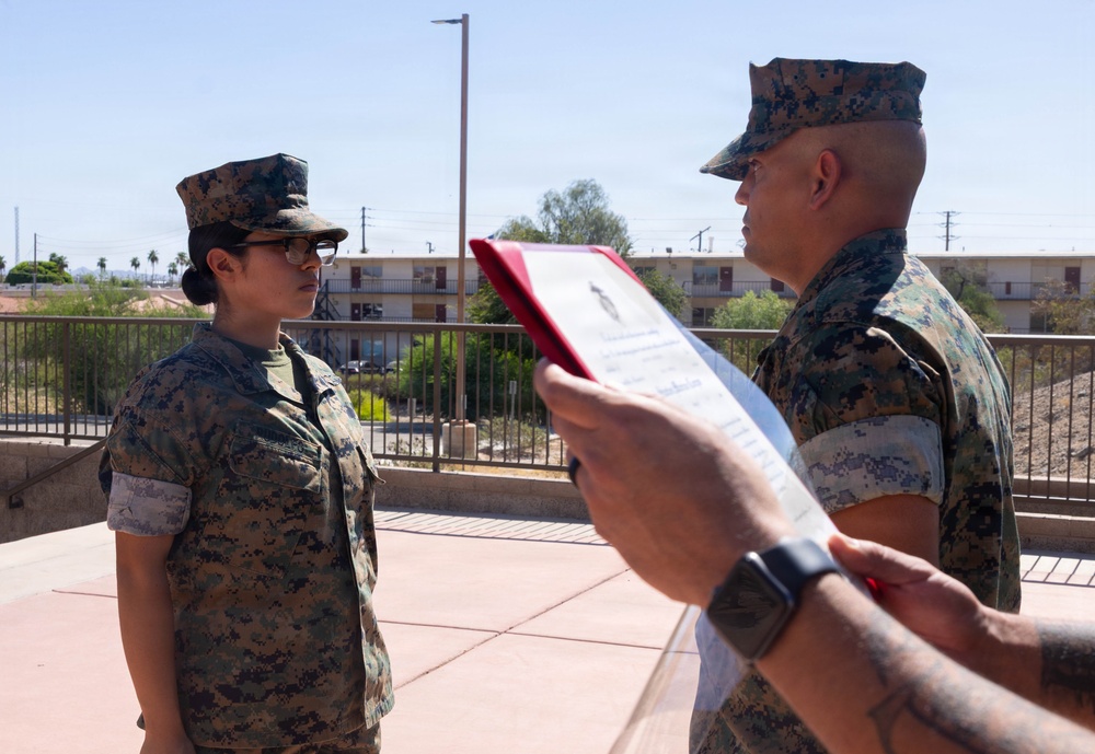 Two U.S. Marines in MARPAT digital camouflage uniforms are engaged in what appears to be a formal military ceremony, likely a promotion or award presentation. A senior Marine (male) is reading from a document held in a red folder/binder, while a junior Marine (female) stands at attention. The setting appears to be an outdoor elevated area, possibly a rooftop or elevated walkway, in a warm, arid environment.