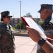 Two U.S. Marines in MARPAT digital camouflage uniforms are engaged in what appears to be a formal military ceremony, likely a promotion or award presentation. A senior Marine (male) is reading from a document held in a red folder/binder, while a junior Marine (female) stands at attention. The setting appears to be an outdoor elevated area, possibly a rooftop or elevated walkway, in a warm, arid environment.