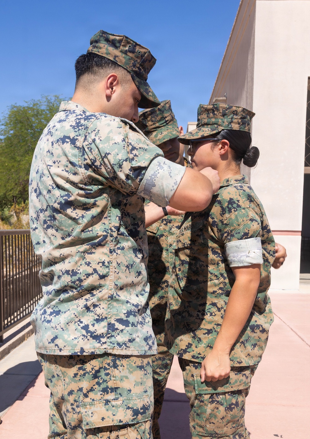 Three military personnel in MARPAT (Marine Pattern) digital camouflage uniforms are gathered outdoors on a paved walkway. One service member appears to be pinning or attaching something to the collar/chest area of a female service member, suggesting a promotion or award ceremony. A third service member is visible in the background observing the event.