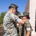 Three military personnel in MARPAT (Marine Pattern) digital camouflage uniforms are gathered outdoors on a paved walkway. One service member appears to be pinning or attaching something to the collar/chest area of a female service member, suggesting a promotion or award ceremony. A third service member is visible in the background observing the event.