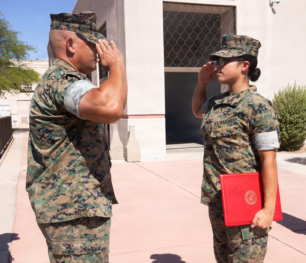 Two U.S. Marines in MARPAT woodland camouflage uniforms are exchanging salutes outdoors at what appears to be a military installation. The female Marine on the right is holding a red ceremonial folder bearing an official seal, suggesting this may be a promotion or award ceremony. The setting is a paved outdoor area adjacent to a tan/beige institutional building.