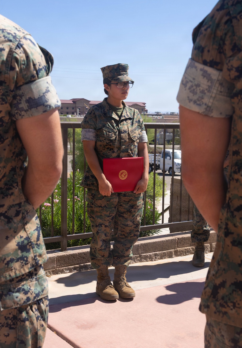 A U.S. Marine in MARPAT camouflage utilities stands at attention on an outdoor walkway or terrace, holding a red presentation folder bearing a gold Marine Corps emblem. The Marine is flanked by two other uniformed personnel visible at the edges of the frame, suggesting a formal military ceremony or award presentation. The setting appears to be a military installation, with base buildings and a vehicle visible in the background.