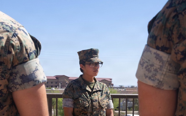 A U.S. Marine in MARPAT camouflage utilities stands at attention on an outdoor walkway or terrace, holding a red presentation folder bearing a gold Marine Corps emblem. The Marine is flanked by two other uniformed personnel visible at the edges of the frame, suggesting a formal military ceremony or award presentation. The setting appears to be a military installation, with base buildings and a vehicle visible in the background.