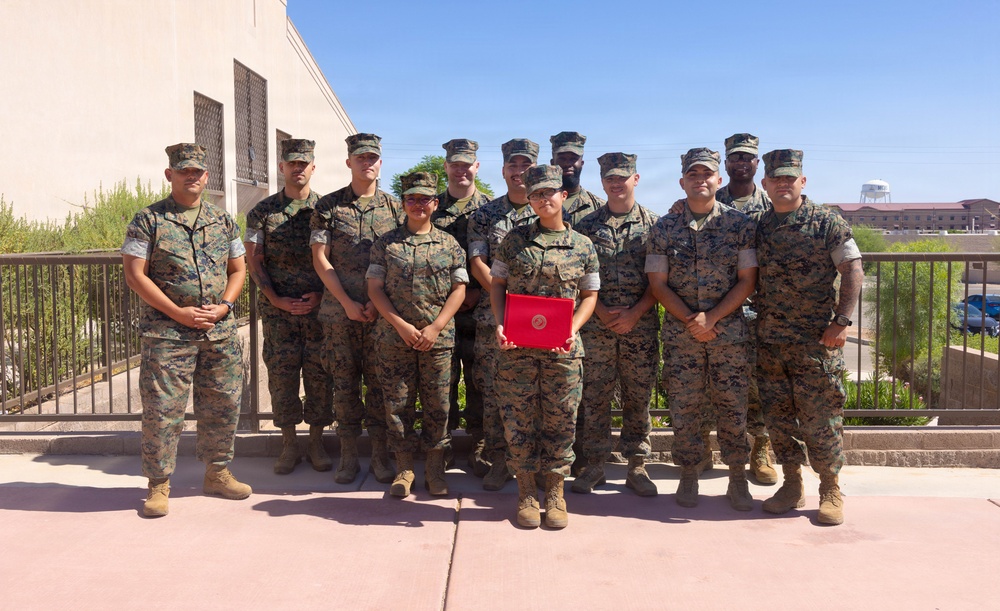 A group of approximately 13 U.S. Marines in woodland MARPAT camouflage uniforms pose together for a formal group photograph outdoors. One Marine in the front center is holding a red folder or certificate, suggesting a recognition or award ceremony. The photo appears to be taken at a military installation, likely in a desert region based on the architecture and landscape.