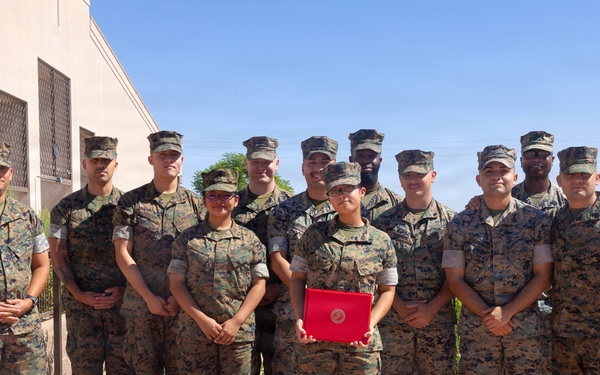 A group of approximately 13 U.S. Marines in woodland MARPAT camouflage uniforms pose together for a formal group photograph outdoors. One Marine in the front center is holding a red folder or certificate, suggesting a recognition or award ceremony. The photo appears to be taken at a military installation, likely in a desert region based on the architecture and landscape.