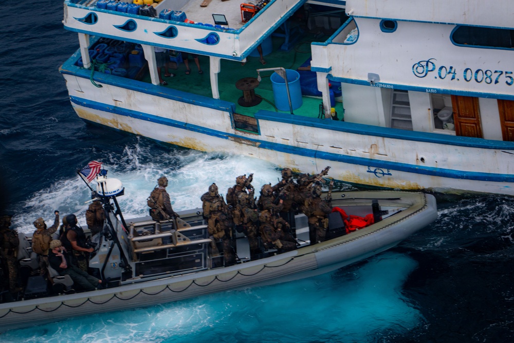 A rigid-hull inflatable boat (RHIB) carrying heavily armed military personnel in full tactical gear is approaching or boarding a larger civilian-style vessel marked 'P.04.00875'. An American flag is visible on the RHIB, indicating U.S. military involvement in what appears to be a maritime interdiction or boarding operation. The larger vessel shows signs of wear and has personnel visible on its upper deck.