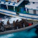 A rigid-hull inflatable boat (RHIB) carrying heavily armed military personnel in full tactical gear is approaching or boarding a larger civilian-style vessel marked 'P.04.00875'. An American flag is visible on the RHIB, indicating U.S. military involvement in what appears to be a maritime interdiction or boarding operation. The larger vessel shows signs of wear and has personnel visible on its upper deck.