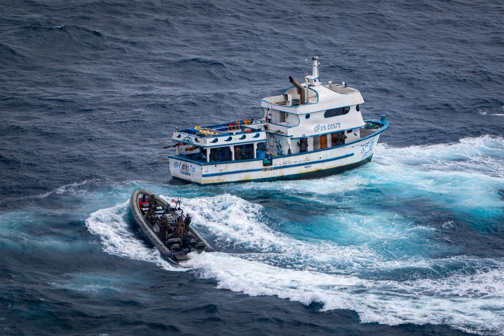 An aerial photograph shows a white and blue fishing vessel named 'Manta' with registration number 'P.C. CC875' being approached by a rigid inflatable boat (RIB) carrying what appear to be armed personnel in dark uniforms. The scene appears to depict a maritime interdiction or law enforcement operation on open ocean waters. Both vessels are underway, with visible wake patterns indicating active movement.