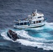 An aerial photograph shows a white and blue fishing vessel named 'Manta' with registration number 'P.C. CC875' being approached by a rigid inflatable boat (RIB) carrying what appear to be armed personnel in dark uniforms. The scene appears to depict a maritime interdiction or law enforcement operation on open ocean waters. Both vessels are underway, with visible wake patterns indicating active movement.