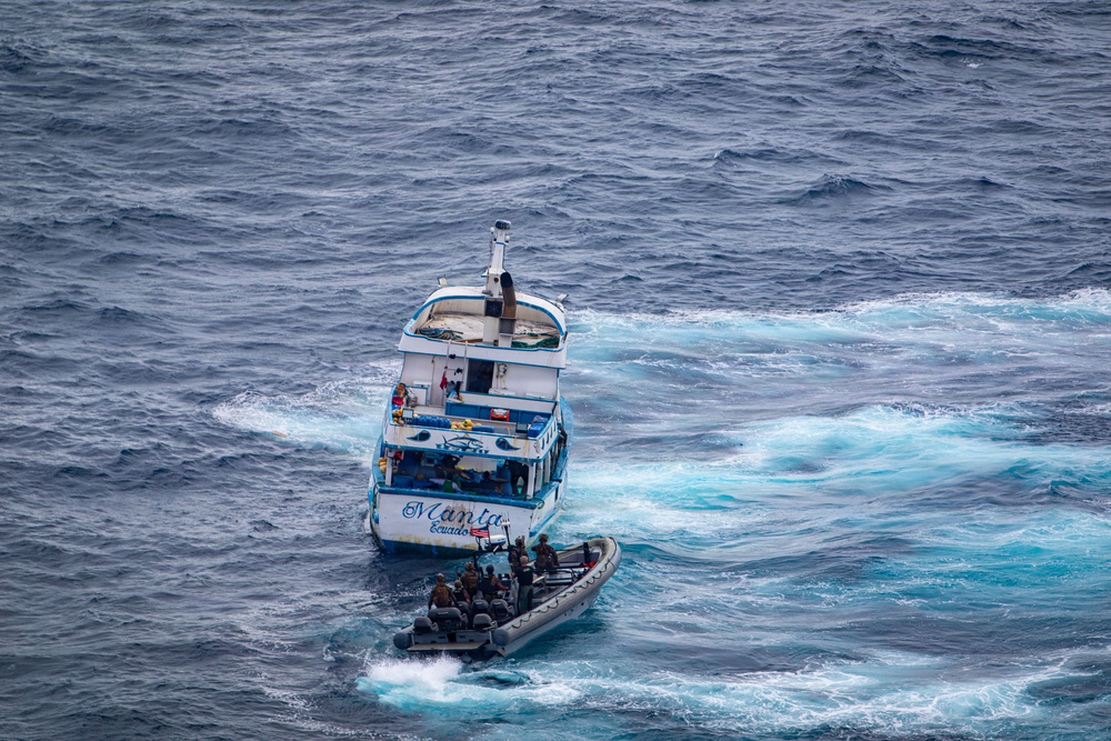 An aerial photograph shows a white and blue fishing vessel named 'Manta, Ecuador' being approached or intercepted by a rigid inflatable boat (RIB) carrying several personnel in dark tactical gear. The scene appears to be a maritime law enforcement or interdiction operation taking place in open ocean waters. The turbulent wake and churning water suggest both vessels are in motion.