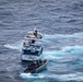 An aerial photograph shows a white and blue fishing vessel named 'Manta, Ecuador' being approached or intercepted by a rigid inflatable boat (RIB) carrying several personnel in dark tactical gear. The scene appears to be a maritime law enforcement or interdiction operation taking place in open ocean waters. The turbulent wake and churning water suggest both vessels are in motion.