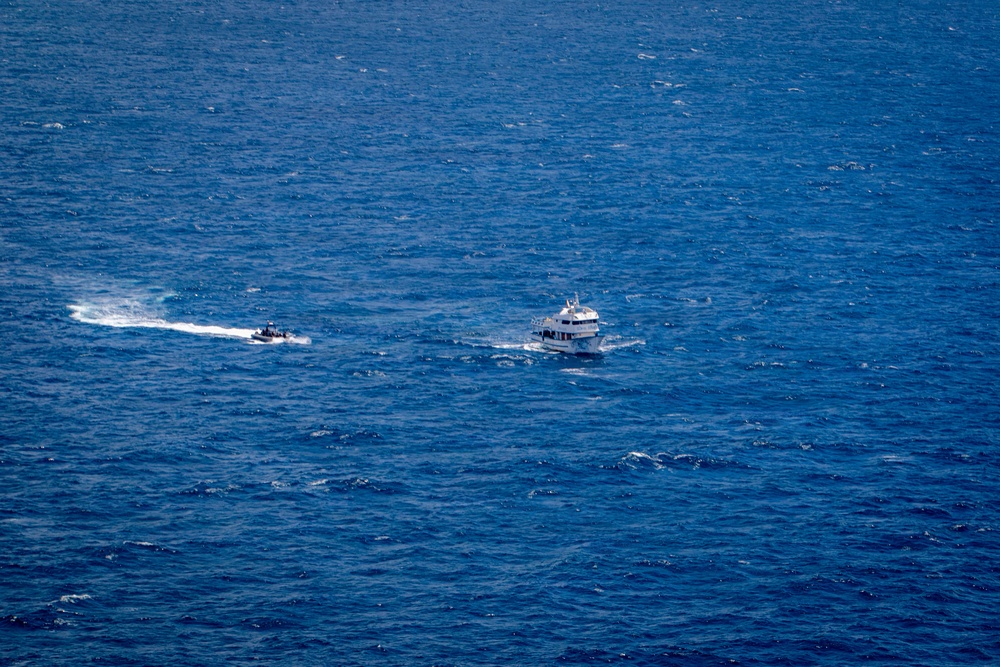 An aerial or elevated view of open blue ocean showing two vessels in proximity to each other. One is a larger white multi-deck vessel, likely a patrol or passenger boat, while the other is a smaller, faster rigid inflatable boat (RIB) leaving a prominent white wake. The scene suggests a possible intercept, escort, or patrol operation at sea.
